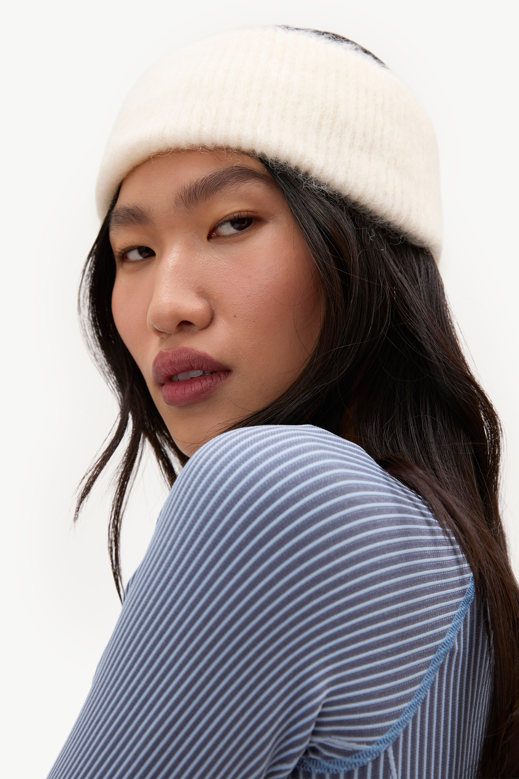 A close-up portrait of a model wearing a cream ribbed knit beanie and a blue and white striped long-sleeve top against a plain white background.