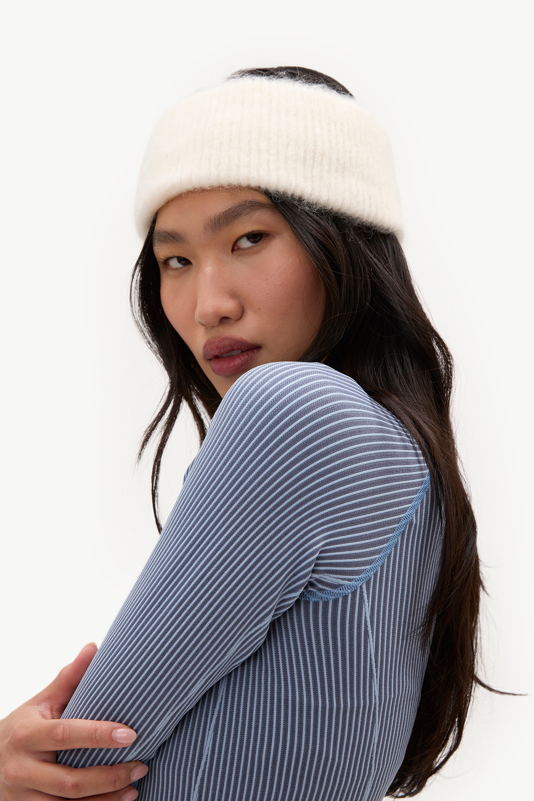 A woman wearing a cream ribbed knit headband and a blue and white vertically striped ribbed top, photographed against a clean white studio background.