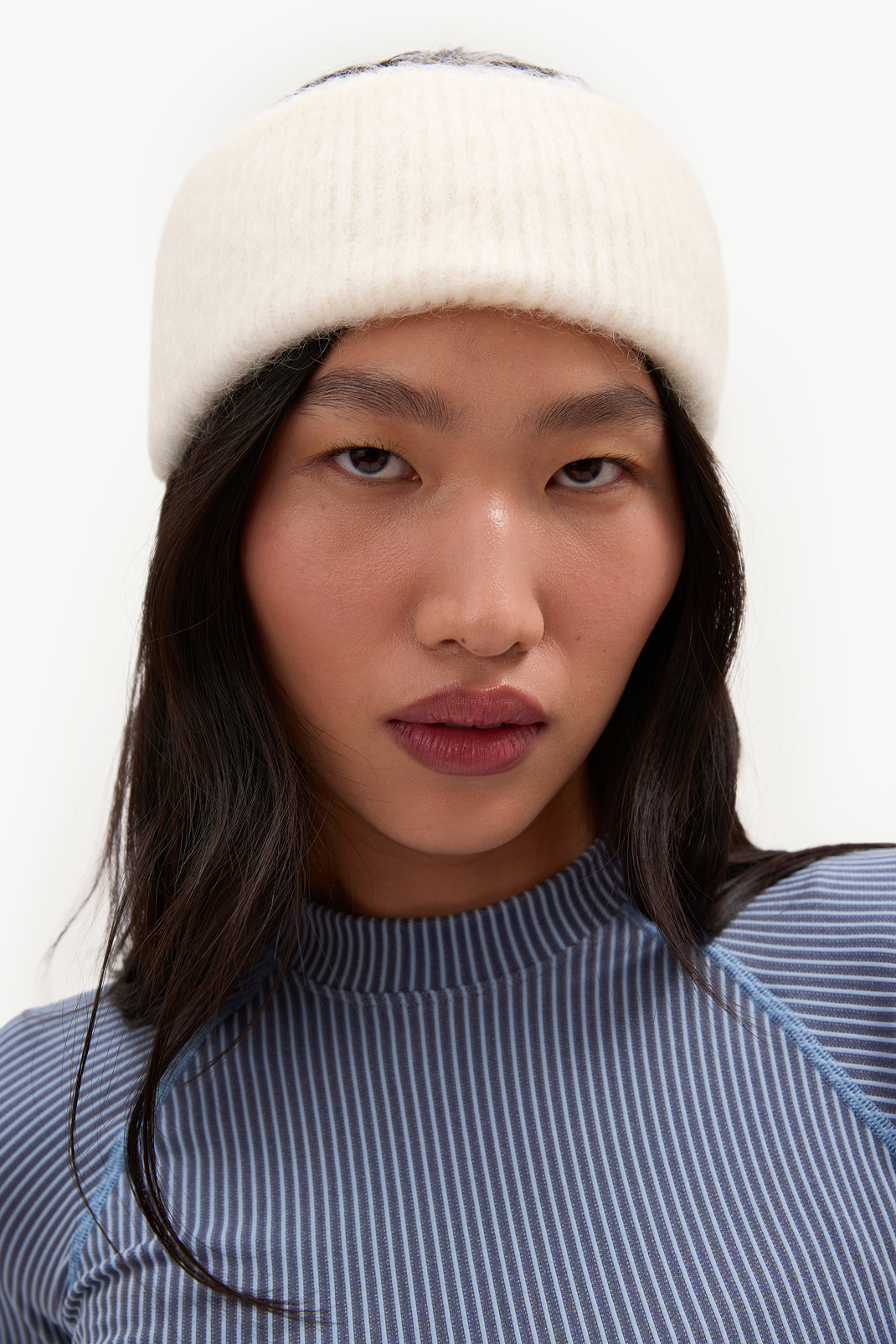 A close-up portrait of a model wearing a cream-colored ribbed knit beanie and a blue and white striped mock neck top against a plain white background.