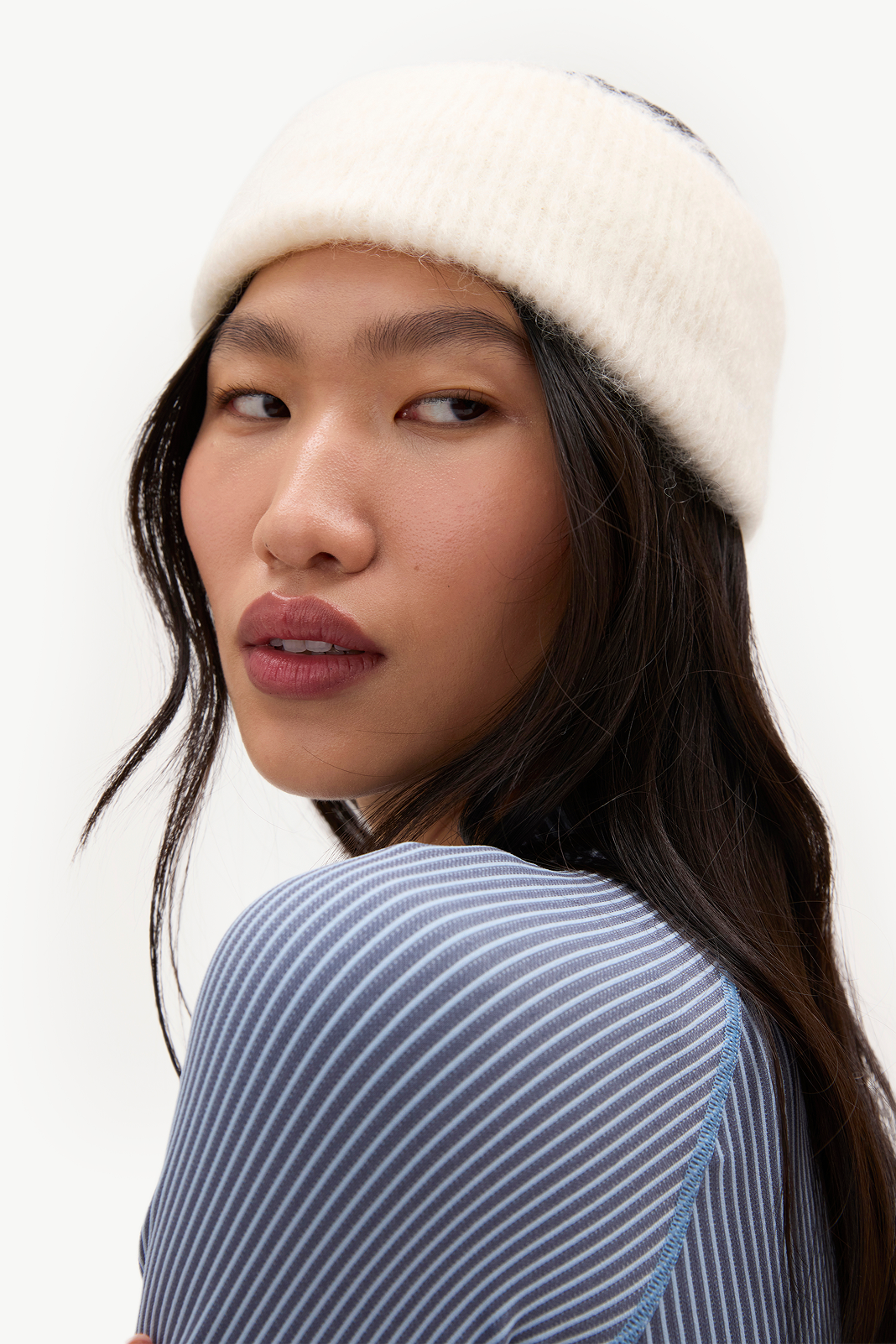 A close-up shot of a model wearing a cream-colored ribbed knit beanie and a blue and white striped ribbed top, photographed against a clean white background.