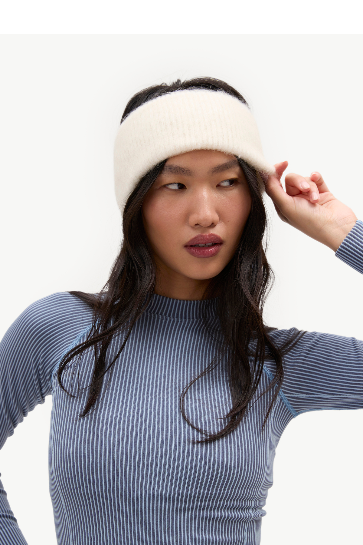 A woman wearing a ribbed cream knit headband and a blue-and-white striped long-sleeve top, photographed against a clean light gray studio background.