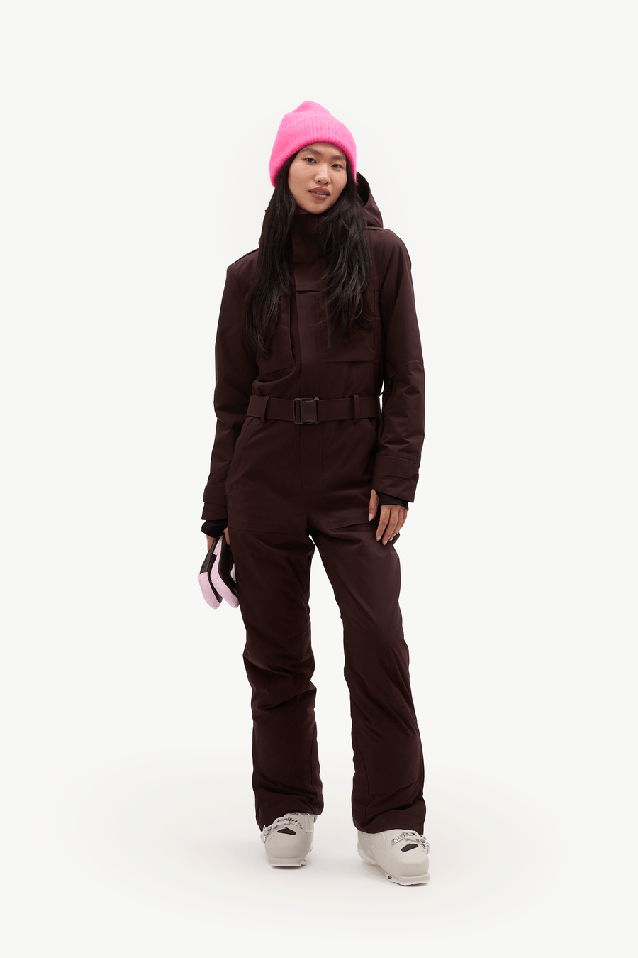 A model stands against a plain white studio background wearing a dark brown ski jacket and matching pants, a bright pink beanie, and white sneakers.