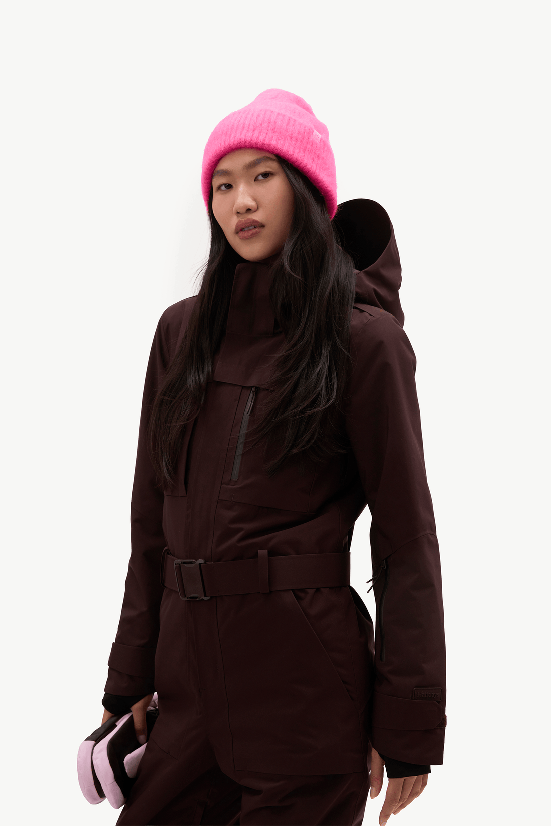 A model wearing a dark brown ski jacket and matching pants, paired with a bright pink knit beanie and holding pink ski goggles, set against a plain light gray background.