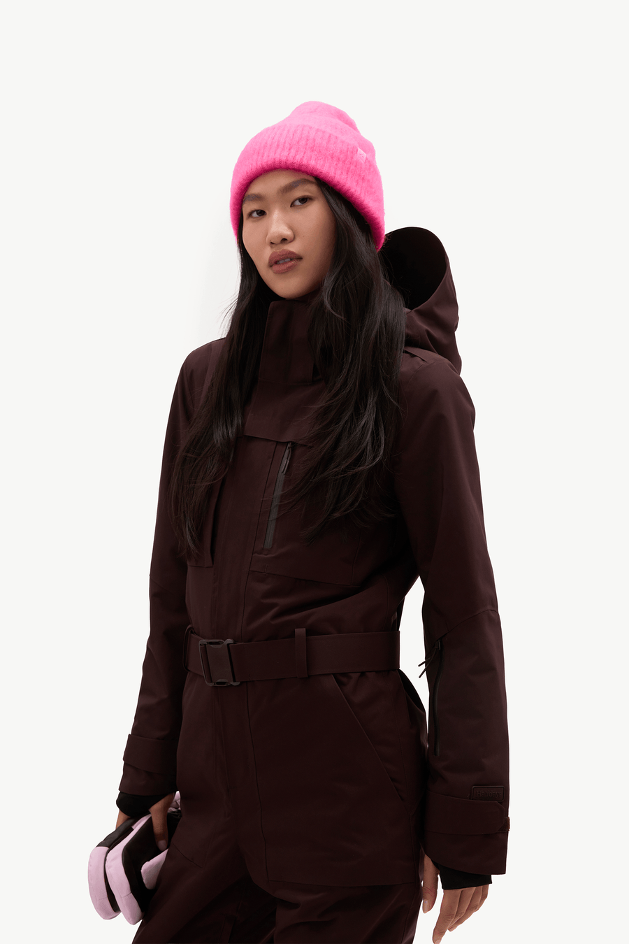 A model wearing a dark brown ski jacket and matching pants, paired with a bright pink knit beanie and holding pink ski goggles, set against a plain light gray background.