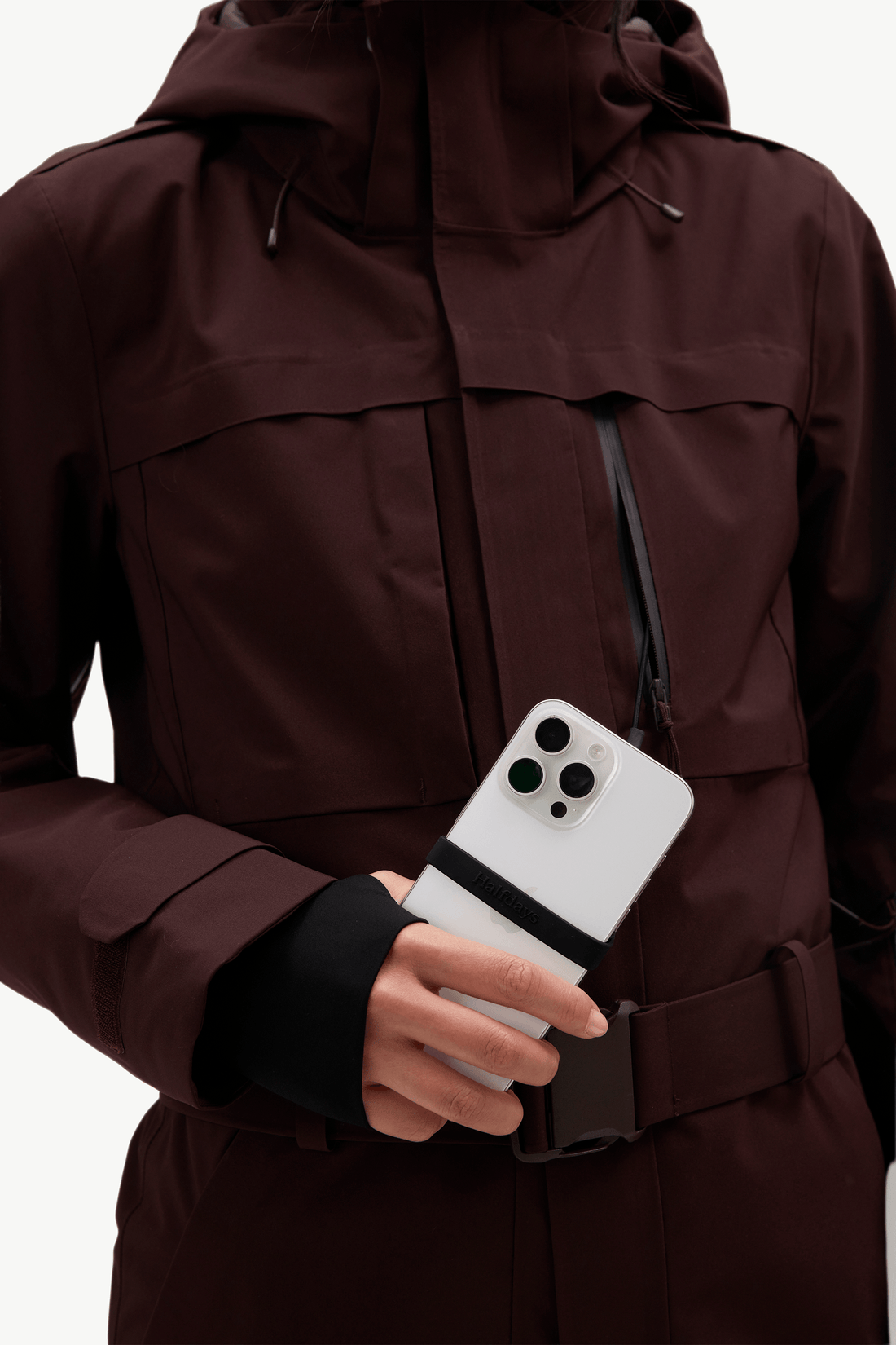 A close-up of a person wearing a dark brown hooded jacket with a black belt and black sleeve detail, holding a white smartphone in their hand.