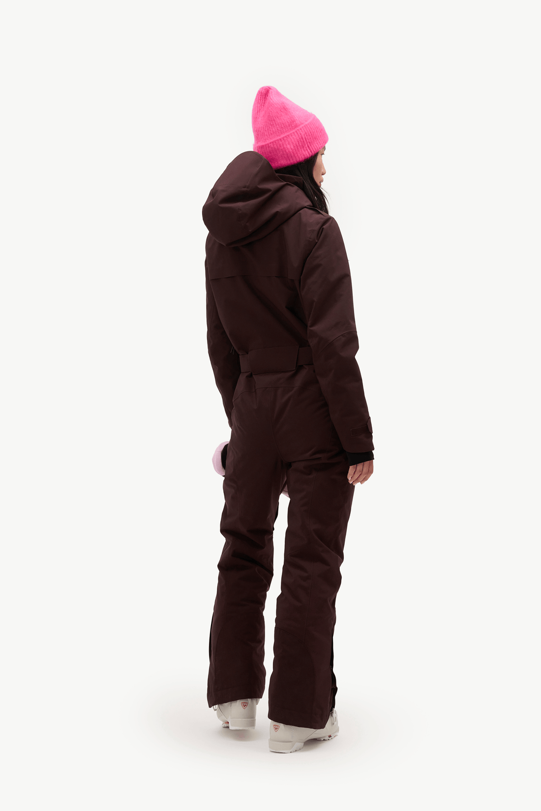 A model shown from the back wearing a dark brown hooded snowsuit, a bright pink knit beanie, and white sneakers, set against a plain white studio background.