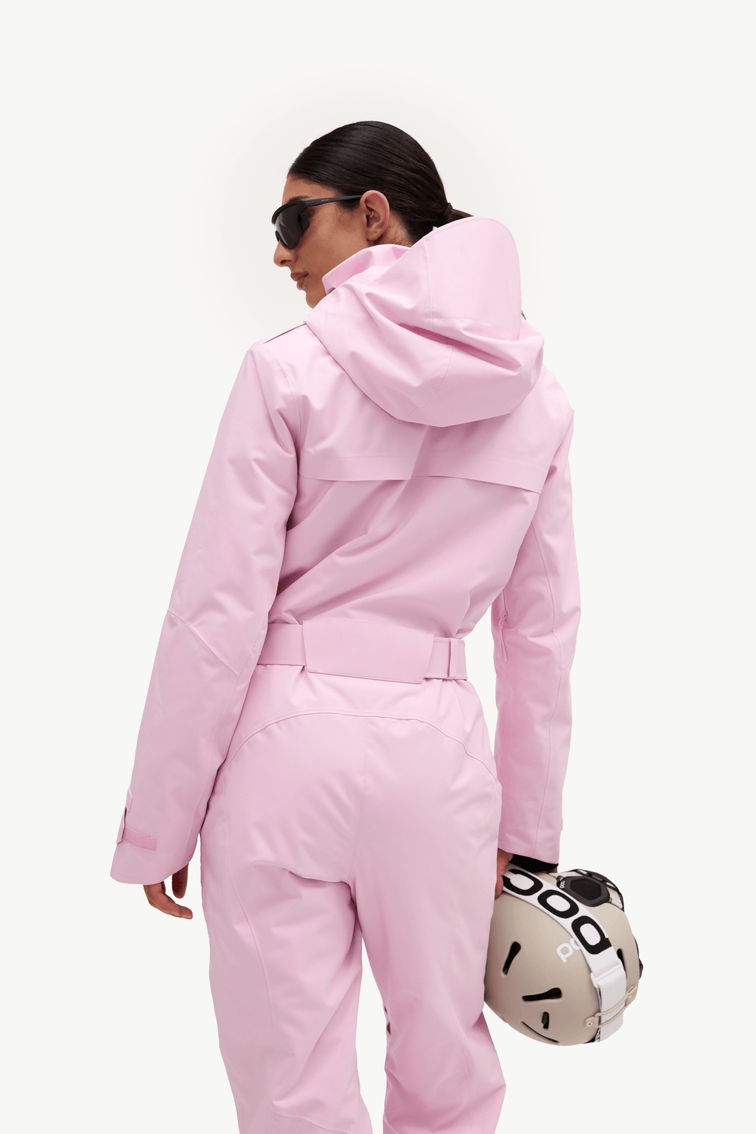 A model from behind wearing a light pink hooded ski jacket and matching pants, holding a patterned ski helmet against a clean white background.