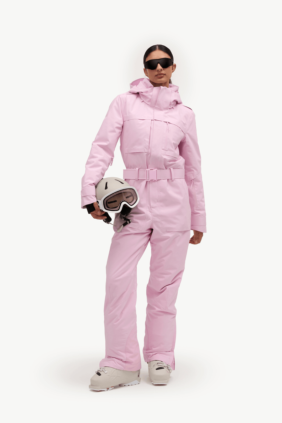 A model wearing a light pink ski suit and holding a white ski helmet with goggles, set against a plain white studio background.