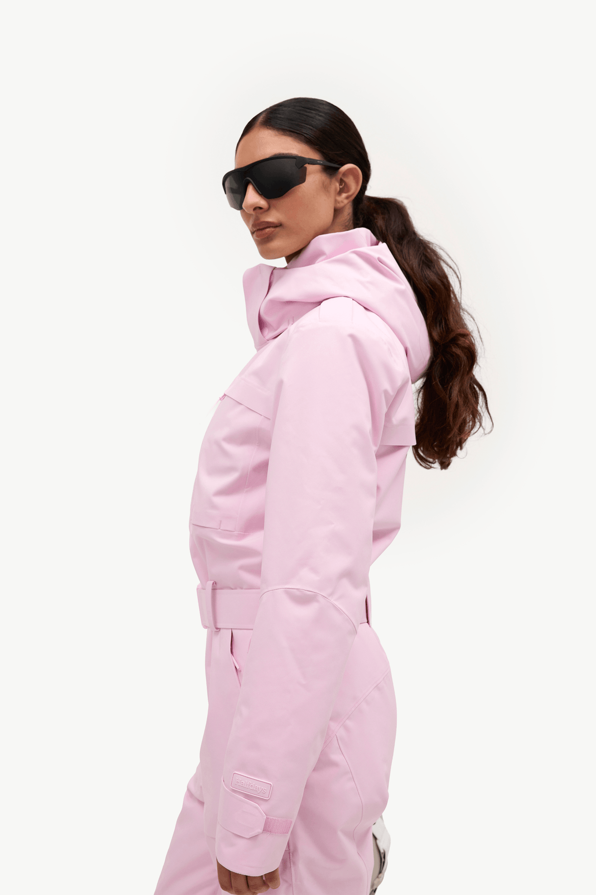 A woman in profile wearing a light pink hooded ski jacket and matching pants, with black sunglasses, against a clean white studio background.