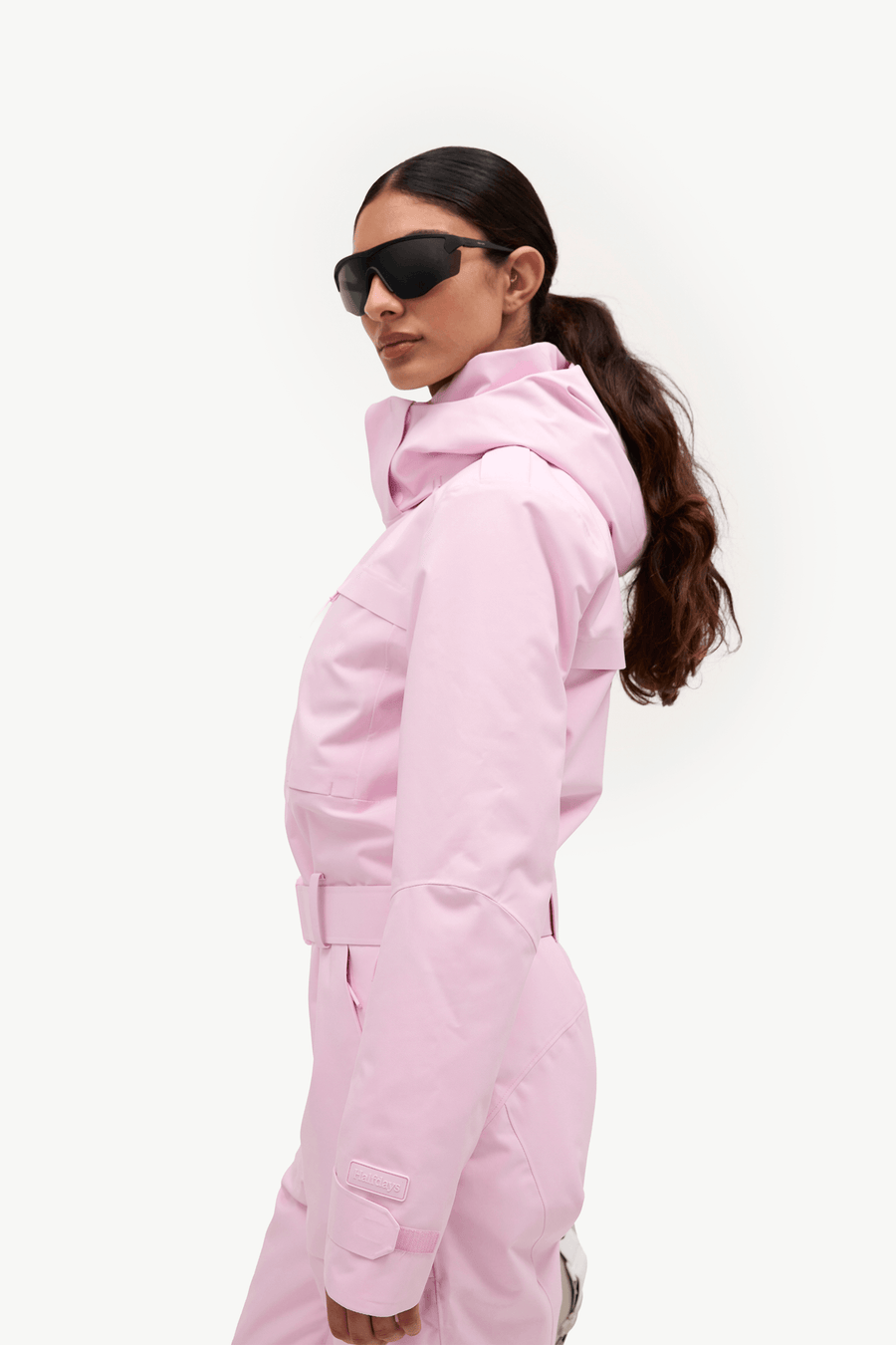A woman in profile wearing a light pink hooded ski jacket and matching pants, with black sunglasses, against a clean white studio background.