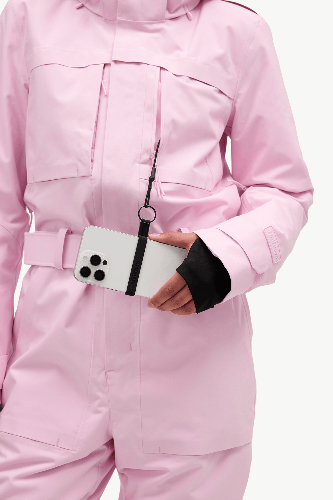 A close-up of a person wearing a light pink technical snowboard jacket, holding a white smartphone in a side pocket. The jacket features a matching pink belt and a black wrist strap accessory attached to the phone.