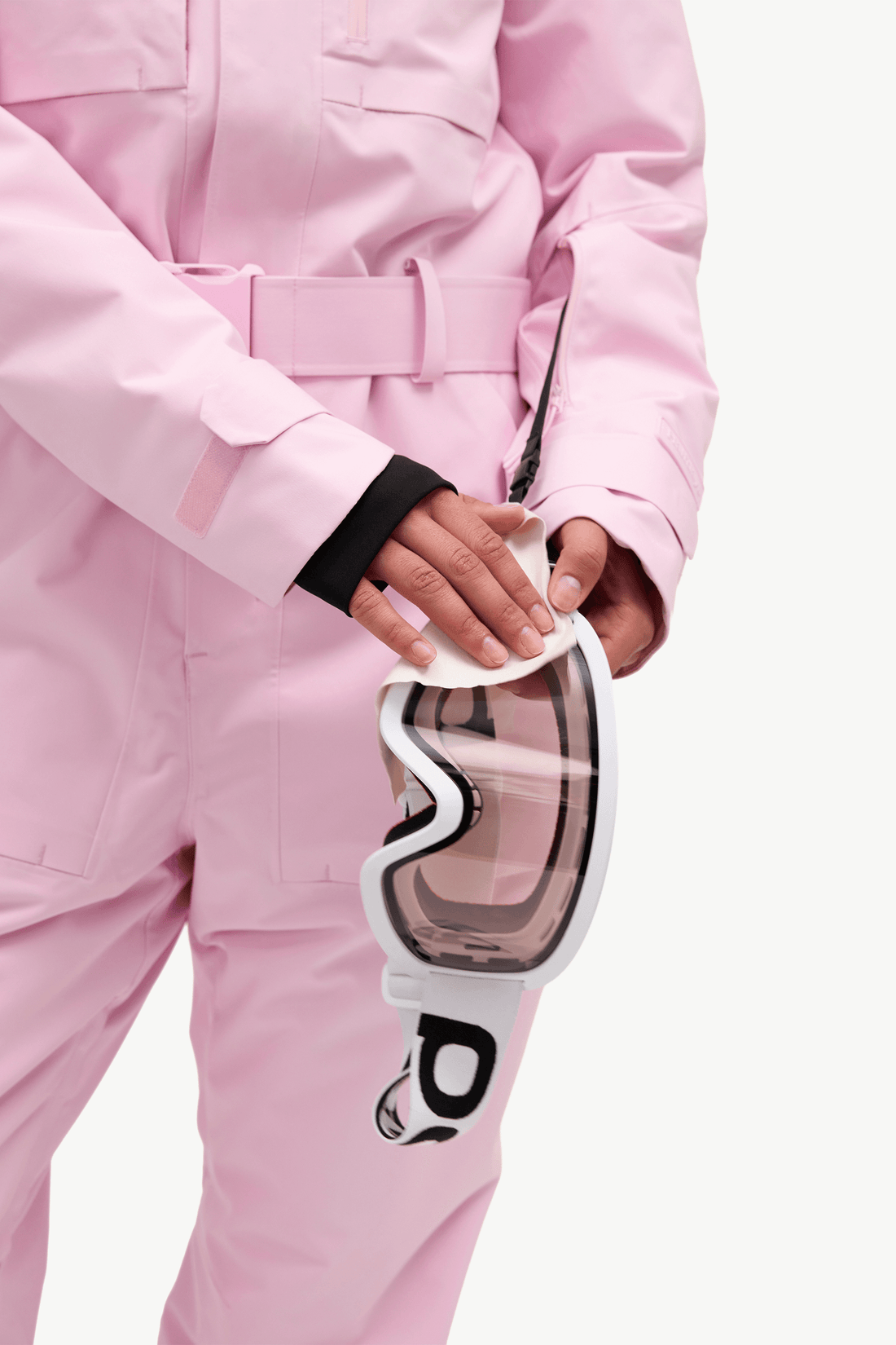 A close-up of a person wearing a light pink snowboard suit and holding a pair of white ski goggles with a clear lens against a clean white background.