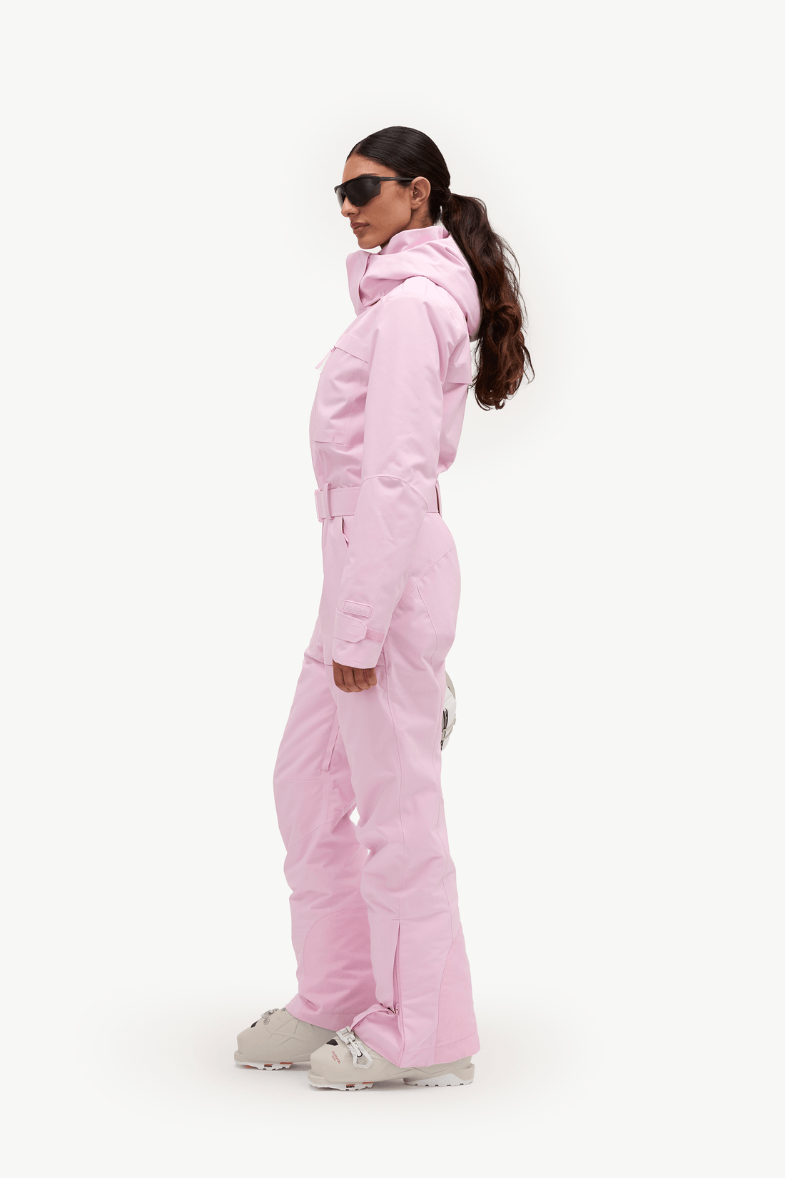 A woman in a light pink ski jacket and matching pants, wearing white ski boots and sunglasses, shown in profile against a plain white studio background.