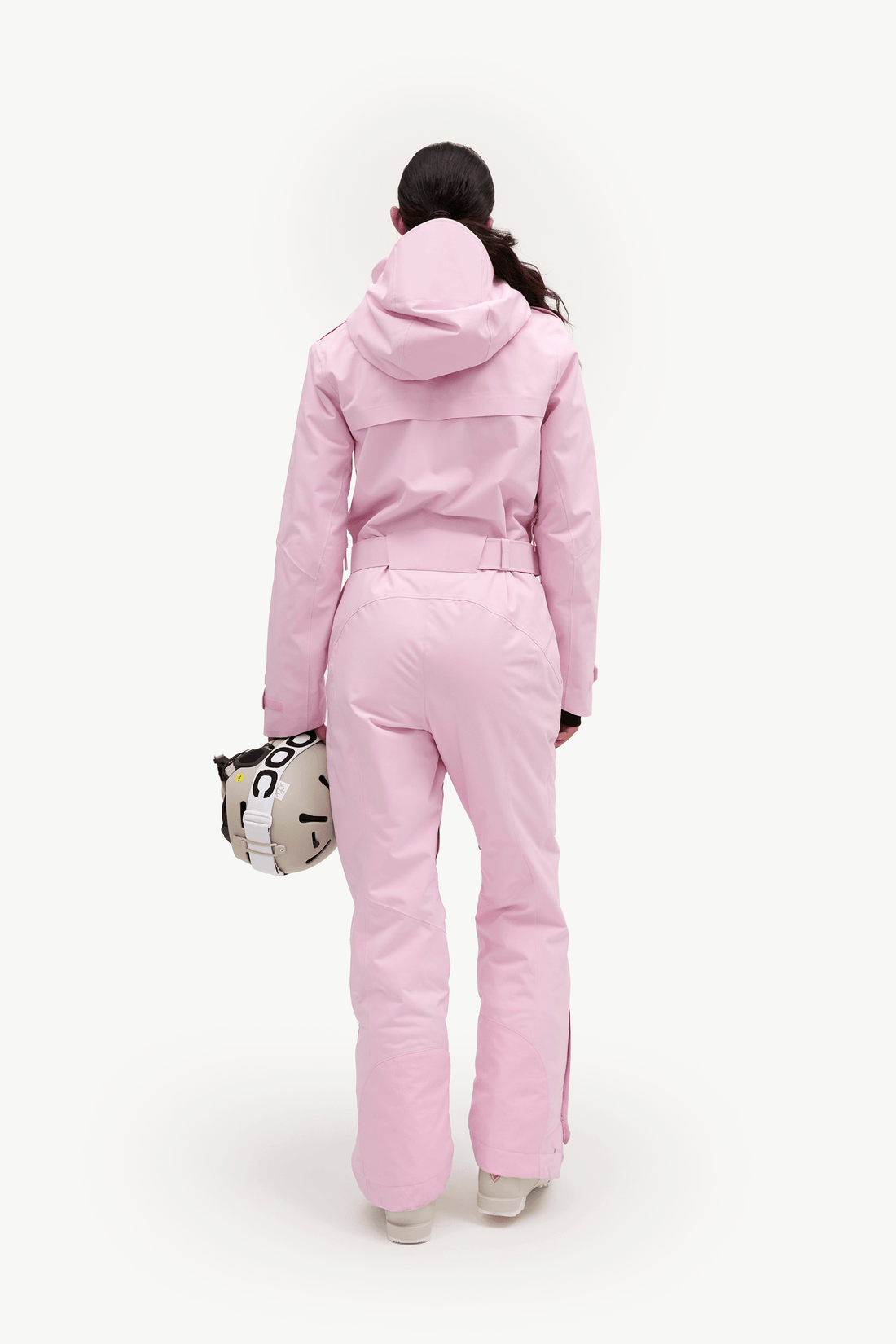 A model viewed from behind wearing a light pink hooded ski jacket and matching ski pants, holding a patterned handbag against a plain white studio background.