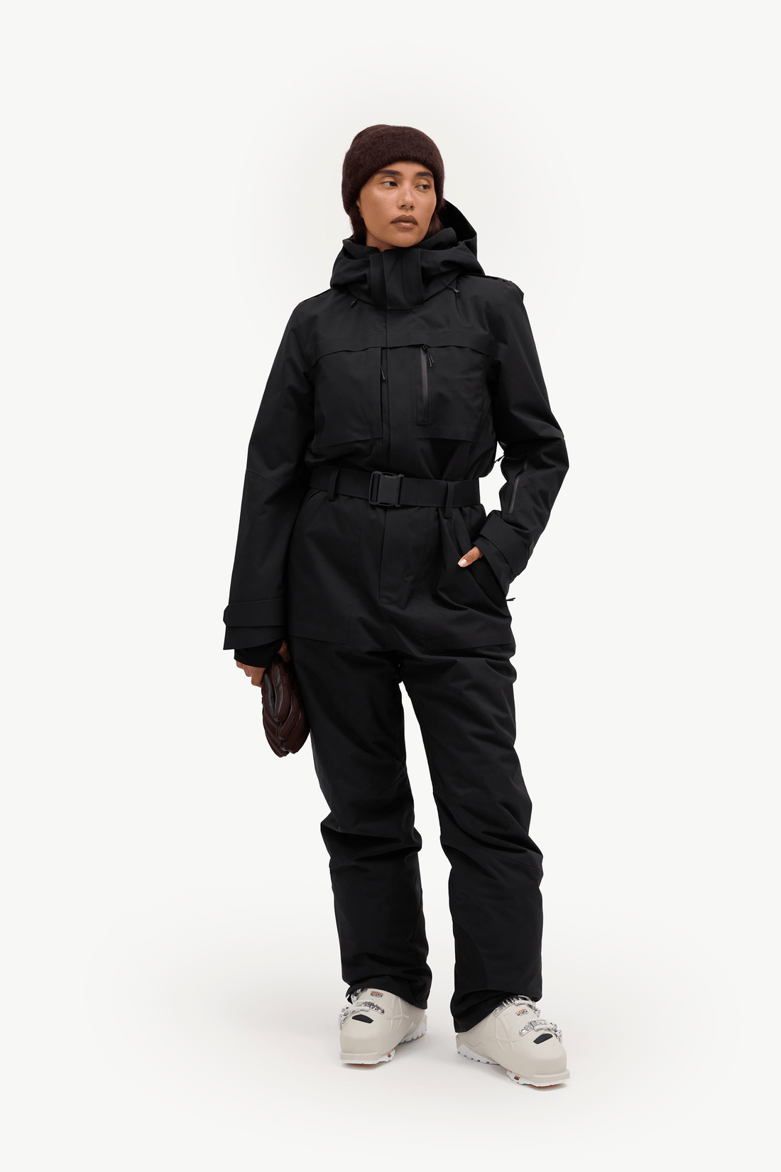 A model wearing a black hooded snowsuit with a matching black beanie and light gray ski boots, standing against a solid white studio background.