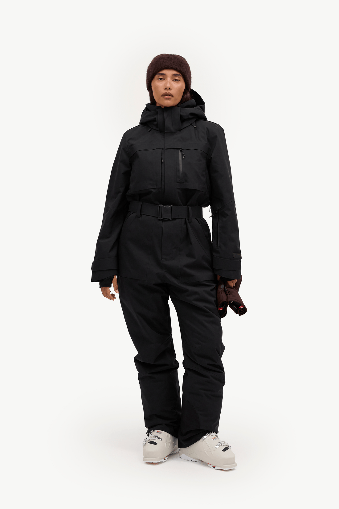 A model stands against a plain white studio background wearing a black hooded ski jacket, matching black ski pants, a dark brown beanie, and light gray snowboard boots.