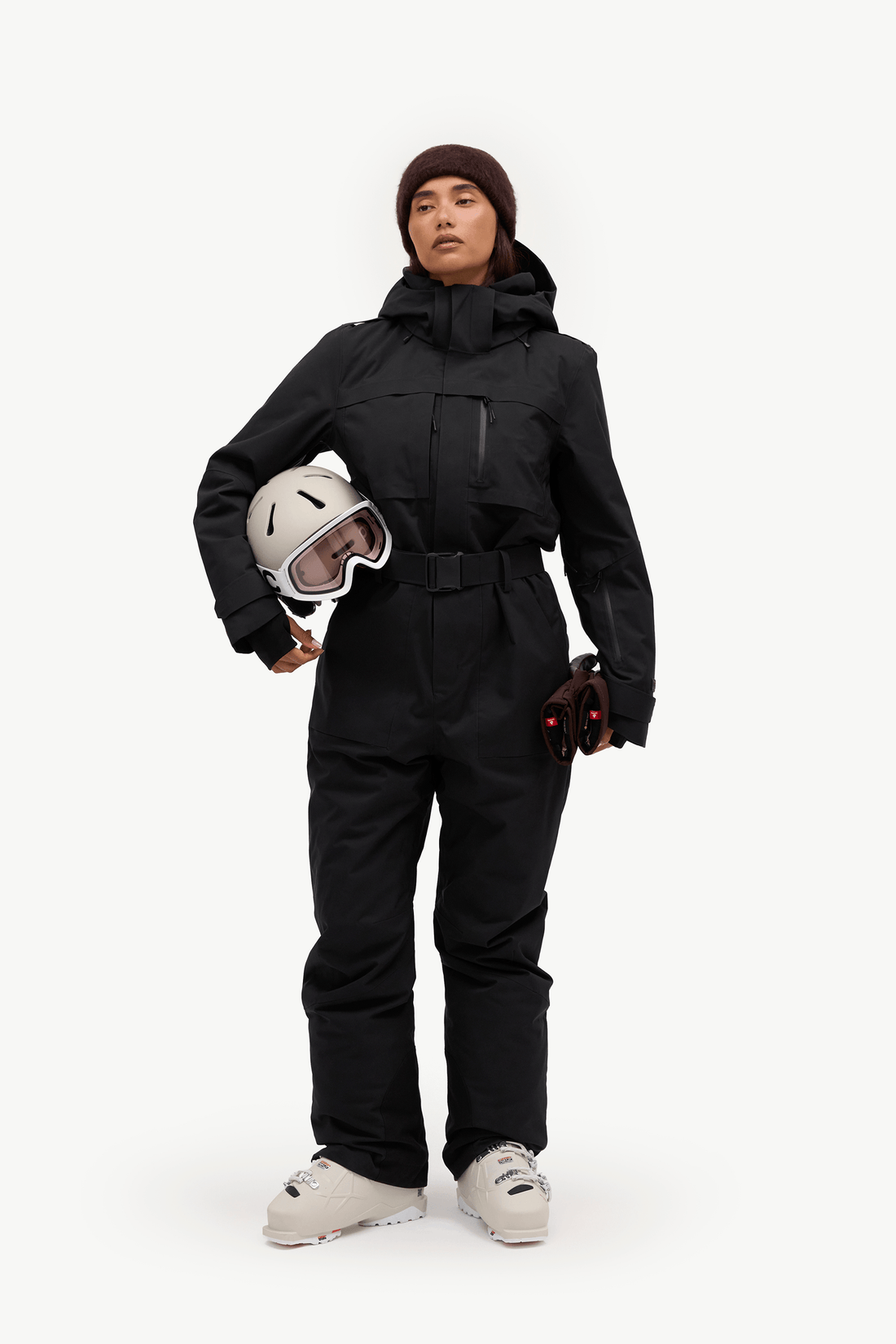 A model wearing a black ski jacket and matching black ski pants, holding a white ski helmet with goggles, standing against a solid white studio background.