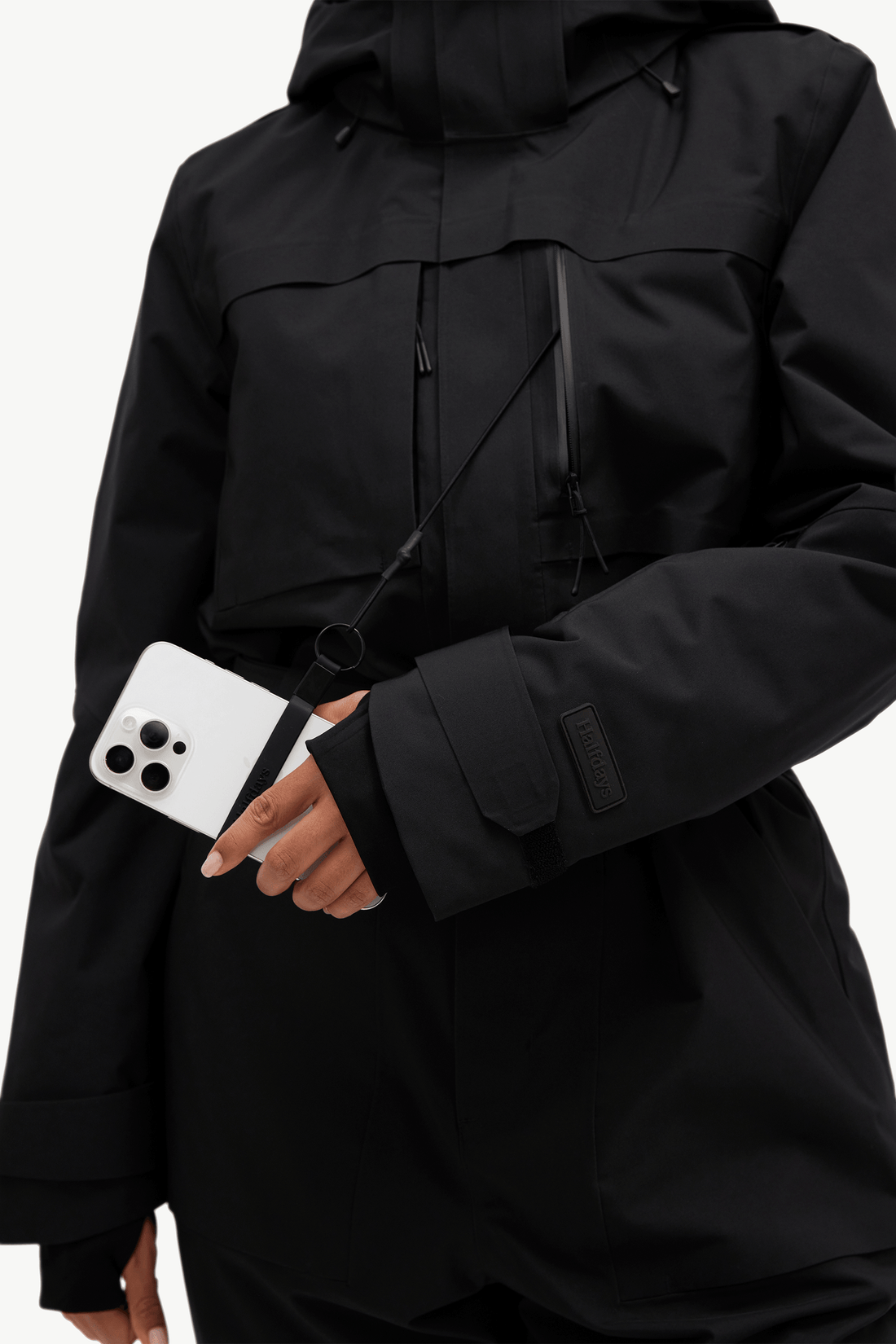 A close-up of a person wearing a black hooded jacket with a black crossbody strap and a white smartphone tucked into the sleeve, against a plain light background.