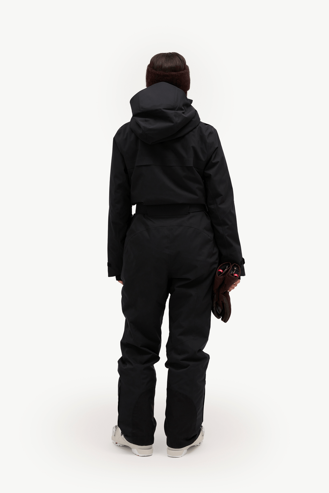 A person viewed from the back wearing a black hooded jacket and matching black pants, standing against a plain light gray studio background. The person is also wearing a dark beanie and holding a dark glove or accessory in one hand.