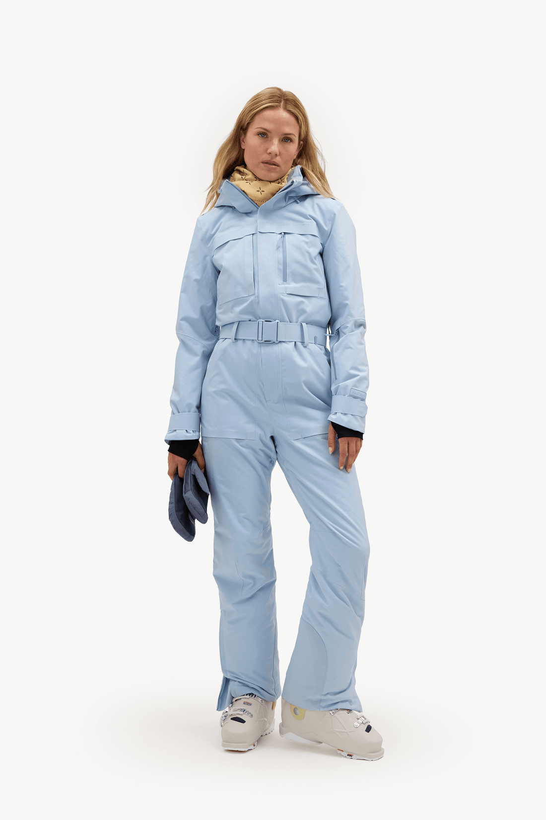 A woman modeling a light blue ski jumpsuit with a matching belt, paired with a yellow neck gaiter, white ski boots, and holding a dark blue glove, against a plain white studio background.