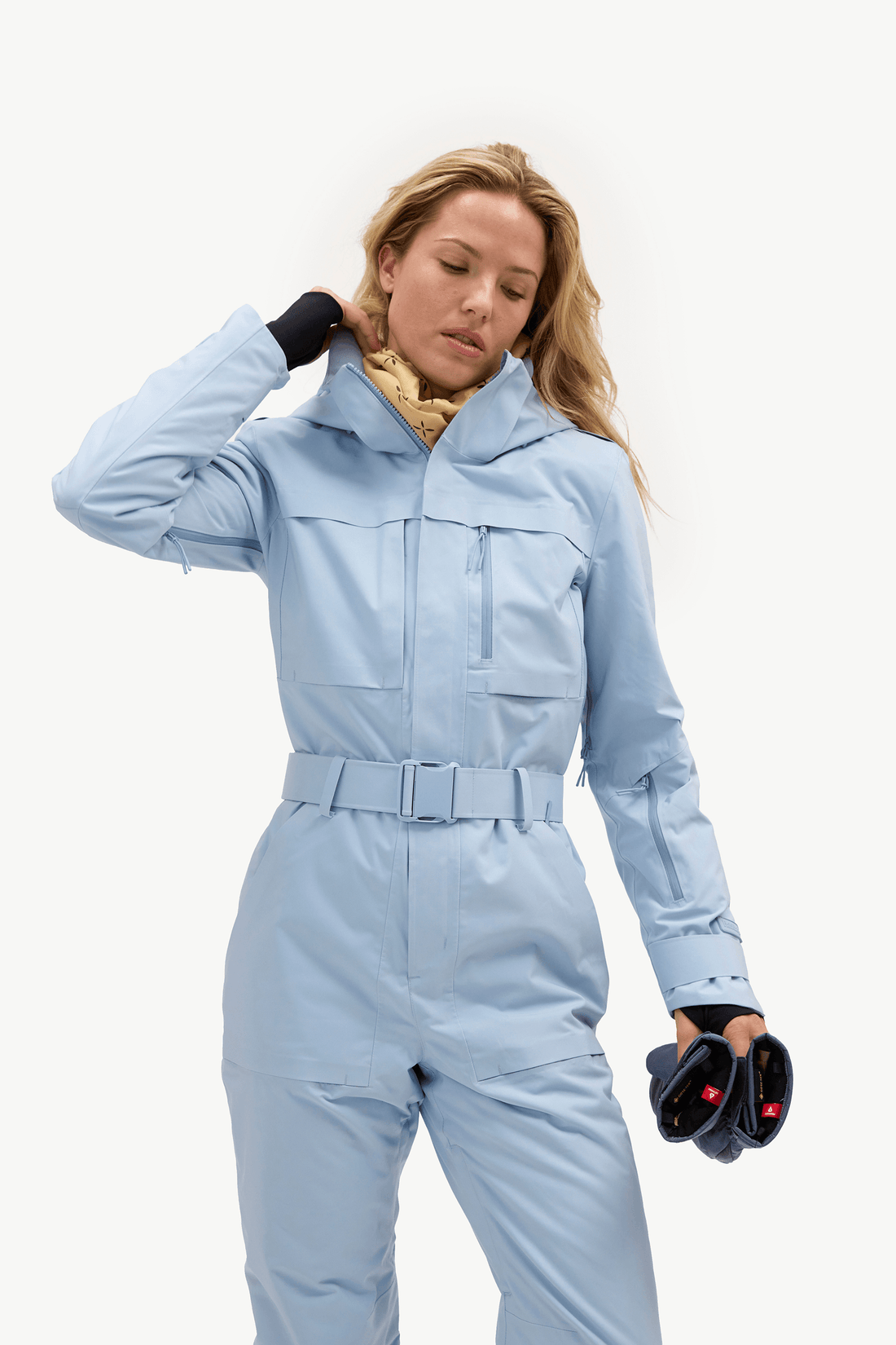 A woman modeling a light blue one-piece snowsuit with a matching belt, black gloves, and a yellow neck gaiter against a plain white studio background.