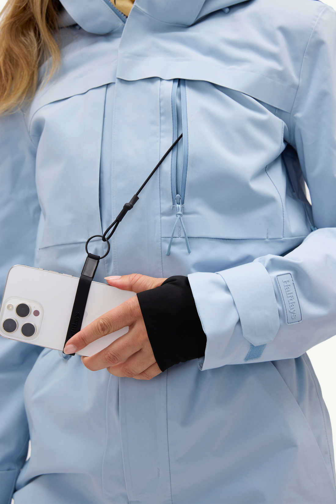 A close-up of a person wearing a light blue technical outdoor jacket, holding a white smartphone with a black wrist strap accessory attached near the zipper.