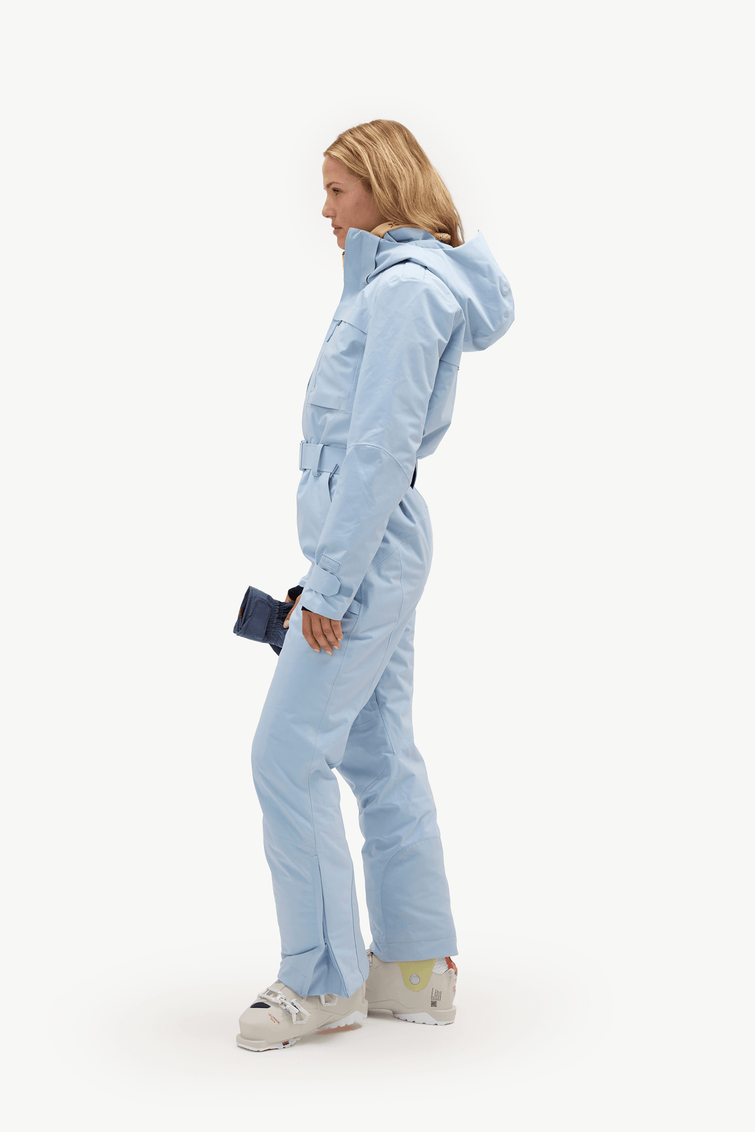 A woman in profile wearing a light blue hooded snowsuit and white ski boots, holding a small dark blue clutch against a clean white studio background.