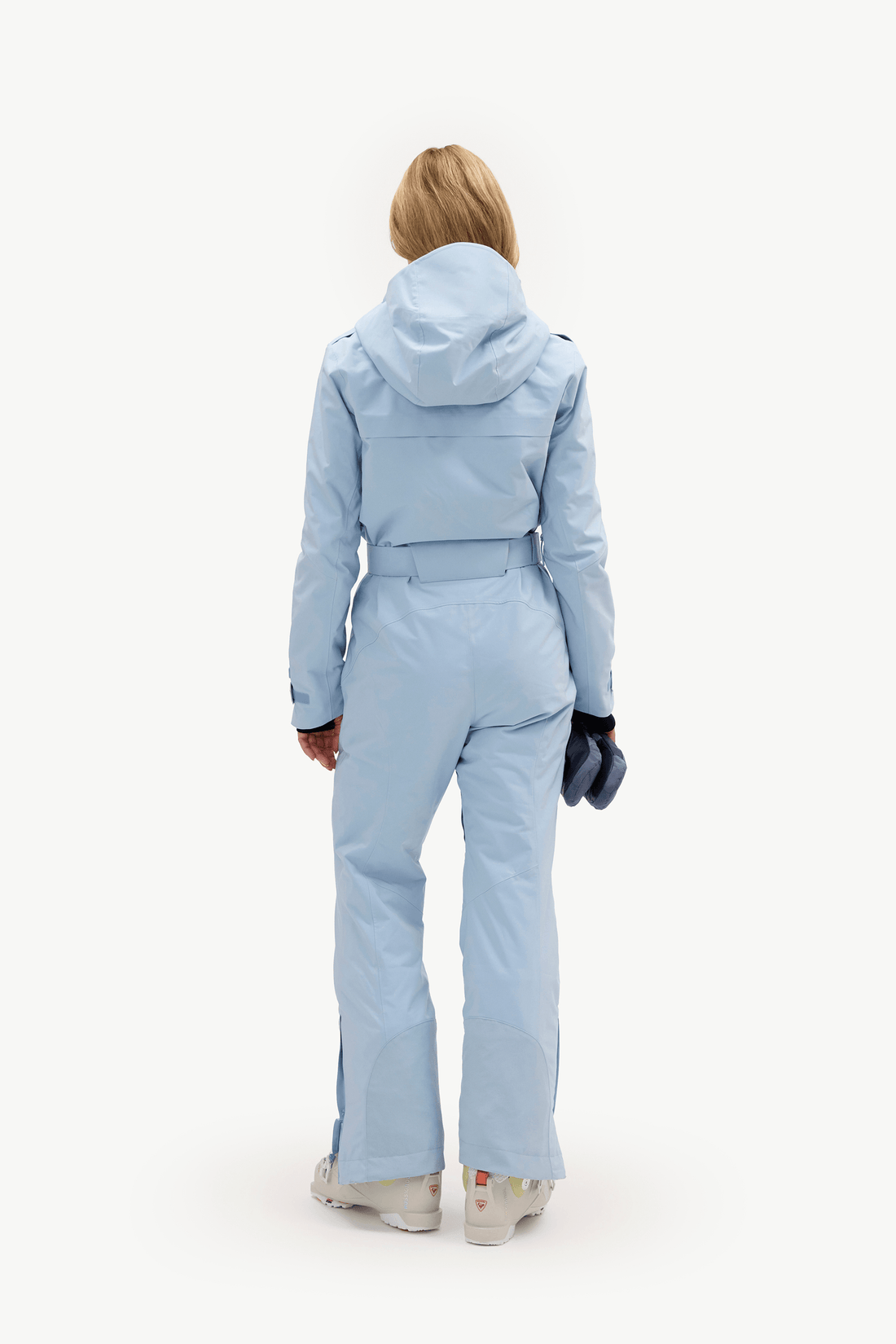A model viewed from the back wearing a light blue hooded ski jumpsuit, holding a dark blue glove, against a solid white background.