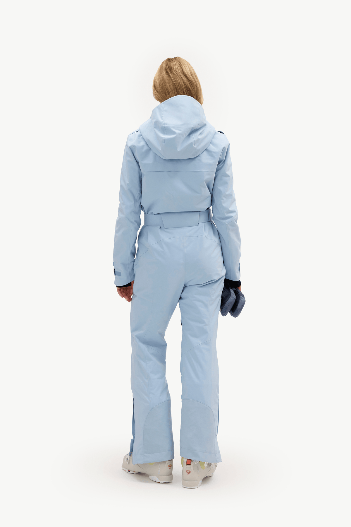 A model viewed from the back wearing a light blue hooded ski jumpsuit, holding a dark blue glove, against a solid white background.