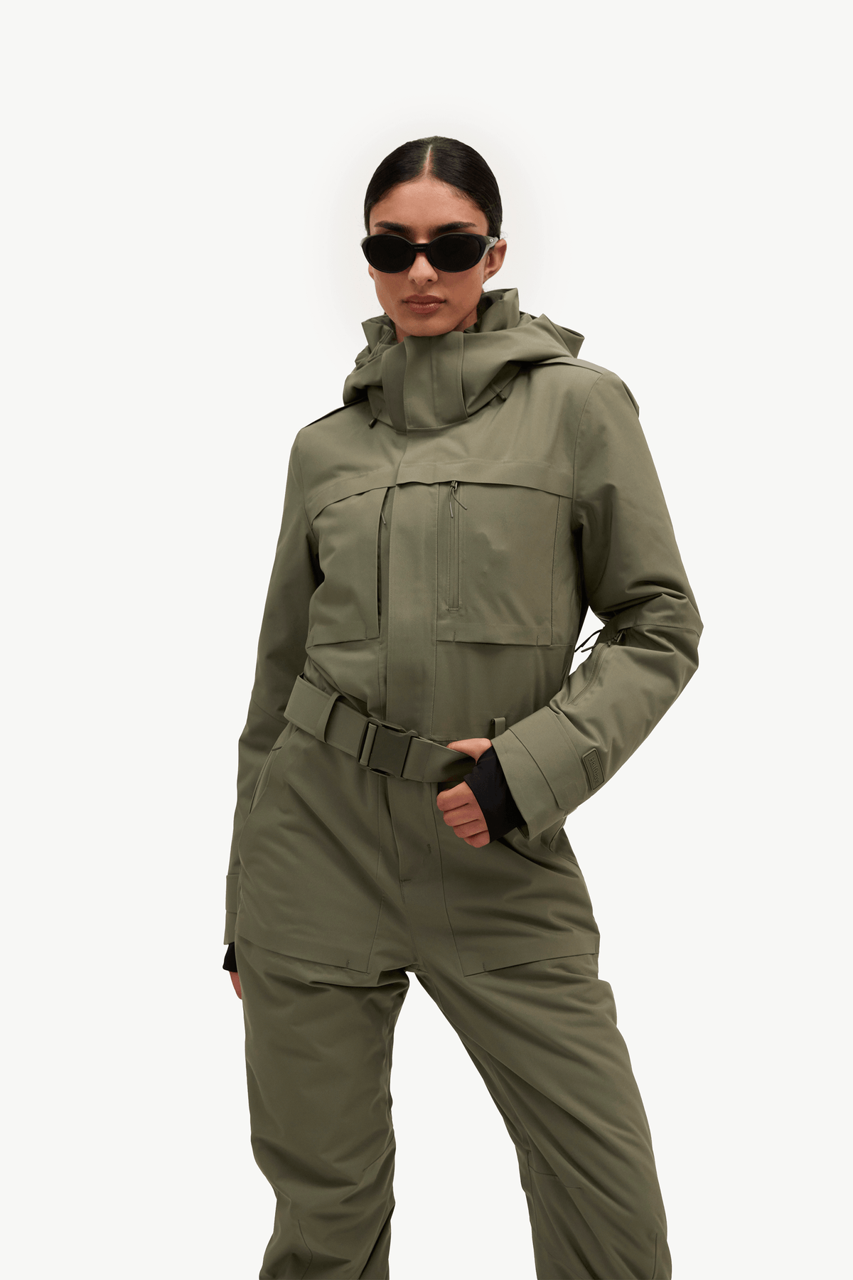 A model wearing a hooded olive green ski jacket and matching insulated pants with a belt, styled with black sunglasses against a plain white studio background.