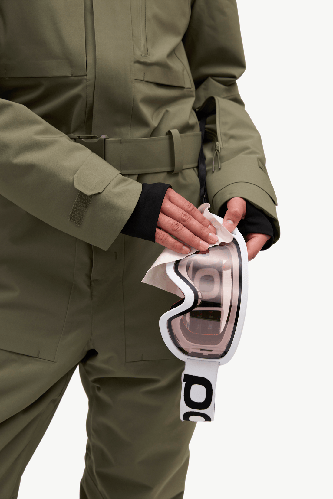 A close-up of a person wearing an olive green snowboard jacket and holding a pair of white ski goggles with a reflective pink lens. The person is shown from the chest down against a clean white background.