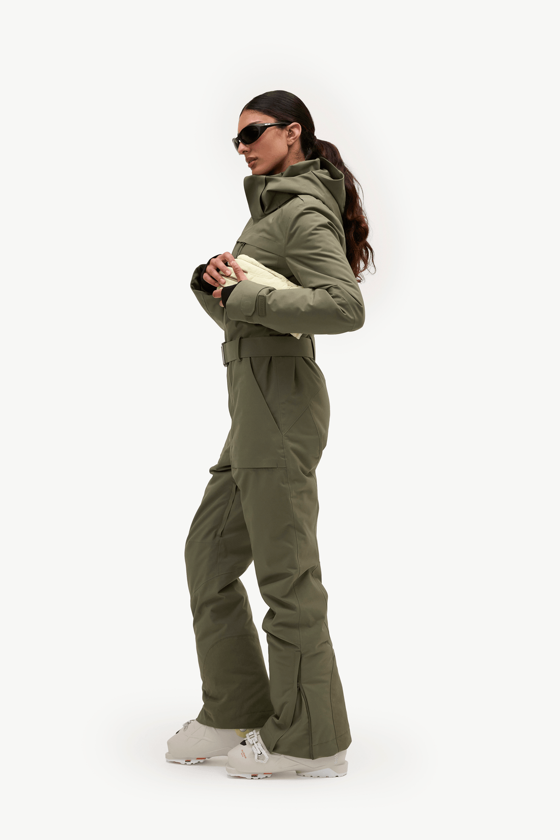 A woman modeling an olive green ski jacket and matching pants set, paired with white sneakers and sunglasses, against a plain white studio background.