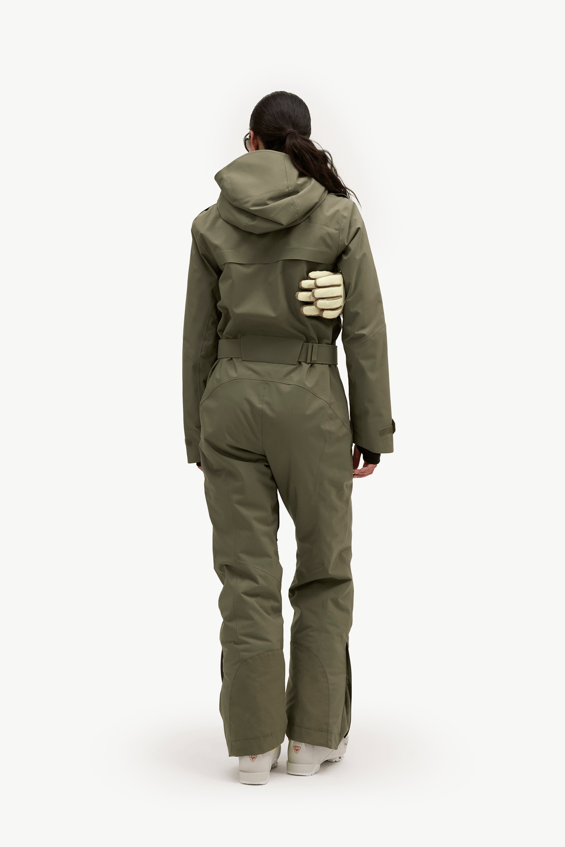 A model viewed from the back wearing an olive green hooded one-piece jumpsuit with integrated gloves and light-colored sneakers, set against a plain white studio background.