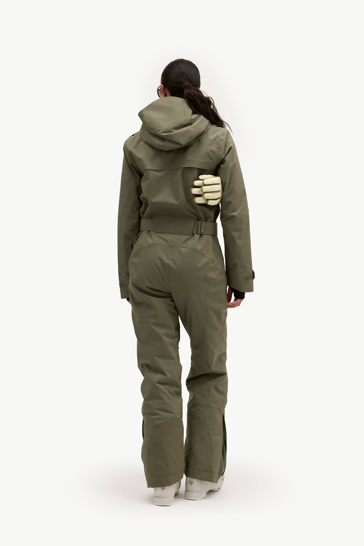 A model viewed from the back wearing an olive green hooded one-piece jumpsuit with integrated gloves and light-colored sneakers, set against a plain white studio background.