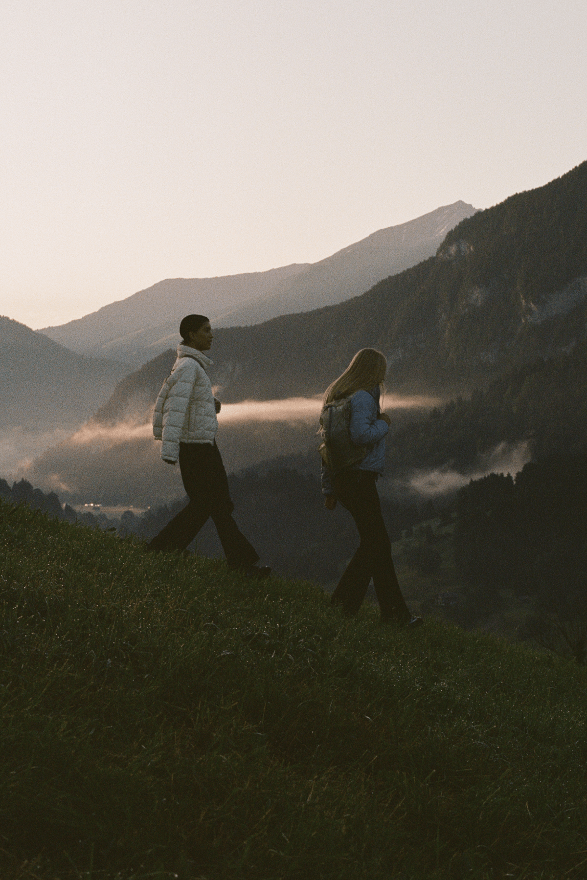 Two people walking side by side on a grassy mountain ridge with misty mountains in the background during golden hour.