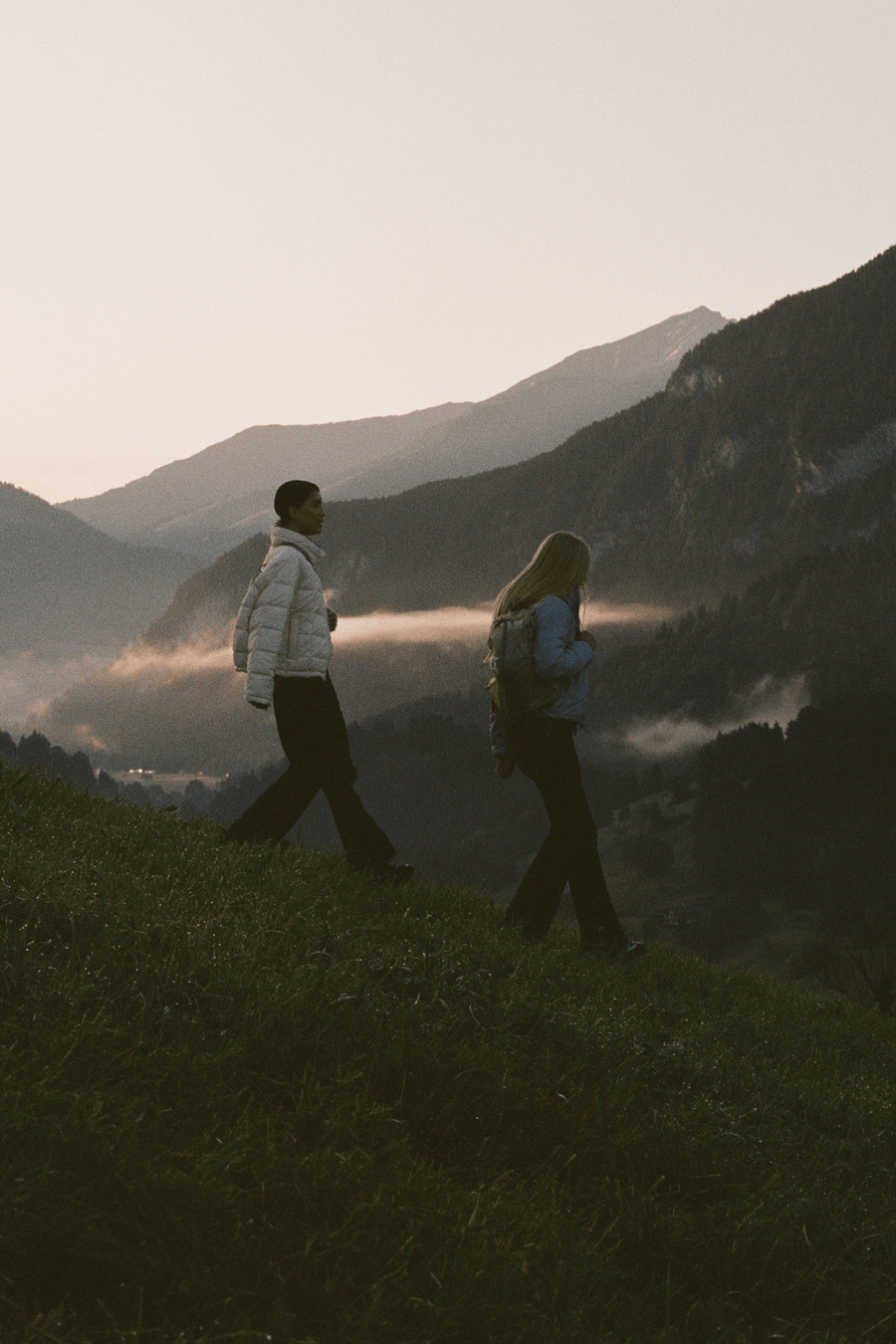 Two people walking side by side on a grassy mountain ridge with misty mountains in the background during golden hour.