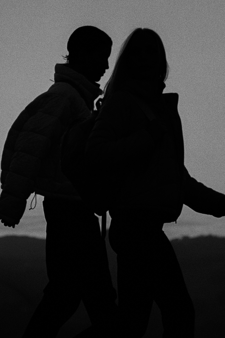 Silhouetted figures of a couple walking side by side against a bright sky, wearing heavy jackets and standing near a railing.