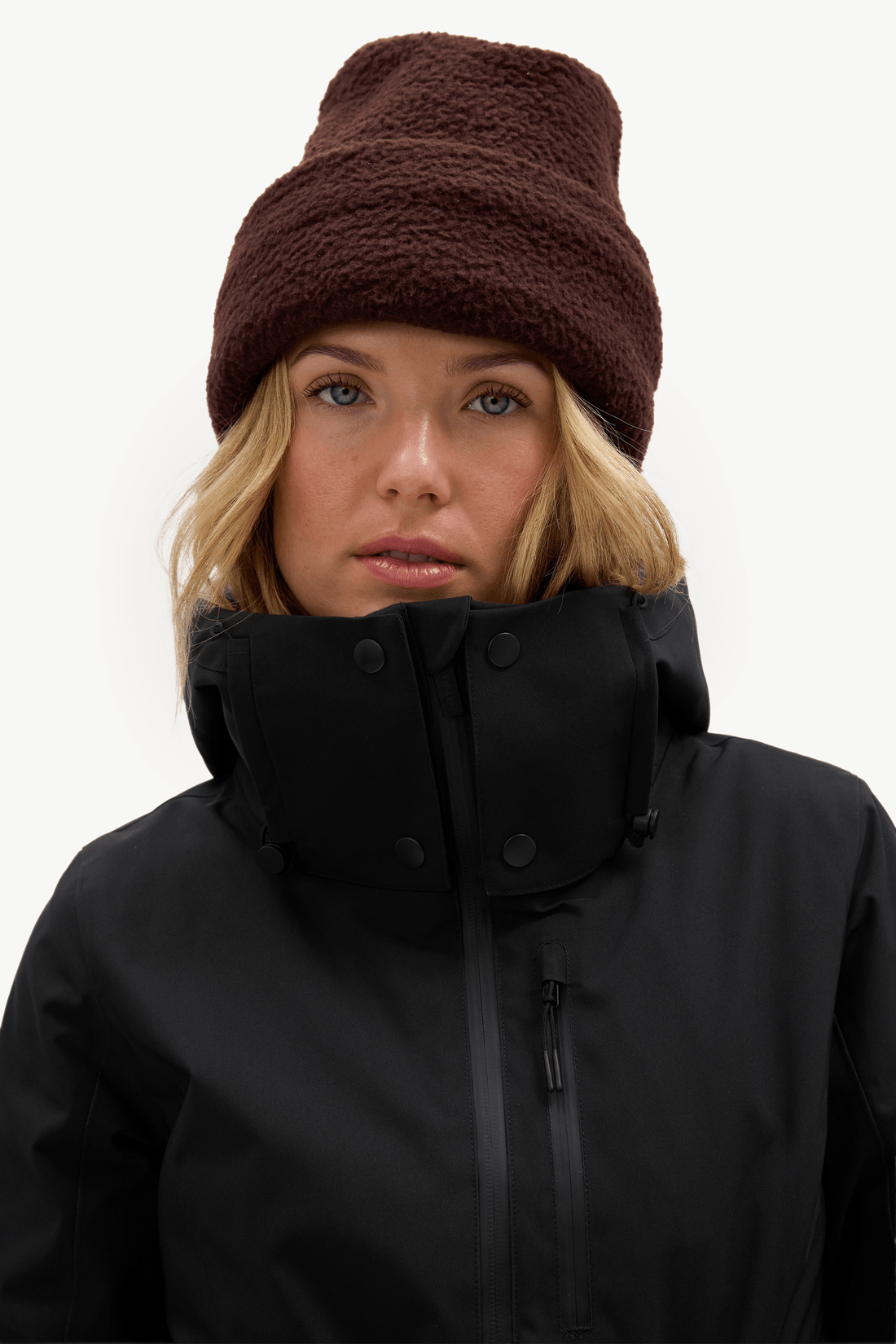 A woman wearing a dark brown fuzzy knit beanie and a black high-collar winter jacket, shown from the chest up against a plain white studio background.