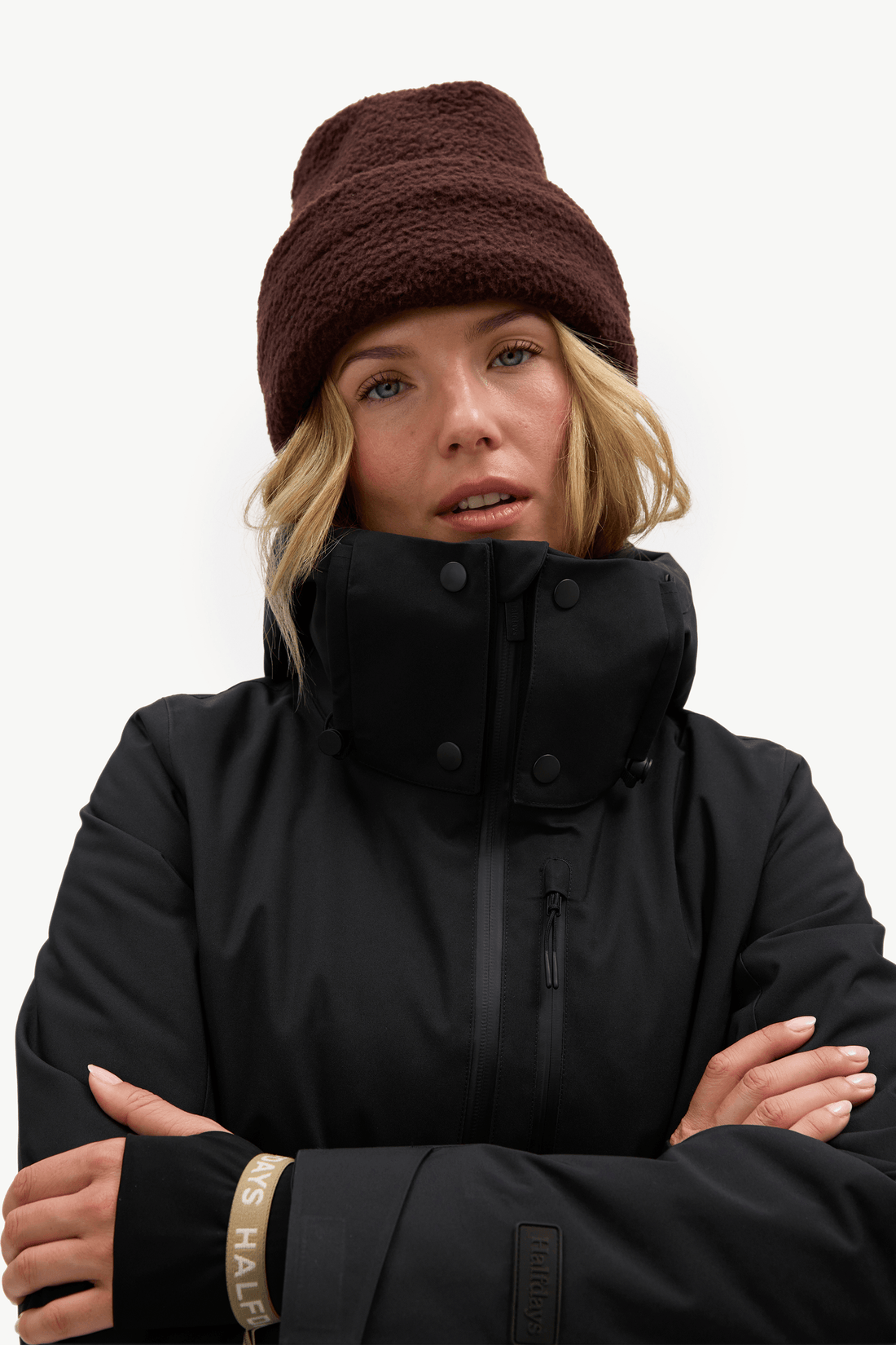 A woman wearing a brown sherpa beanie and a black ski jacket with a high collar, posed against a solid white background.