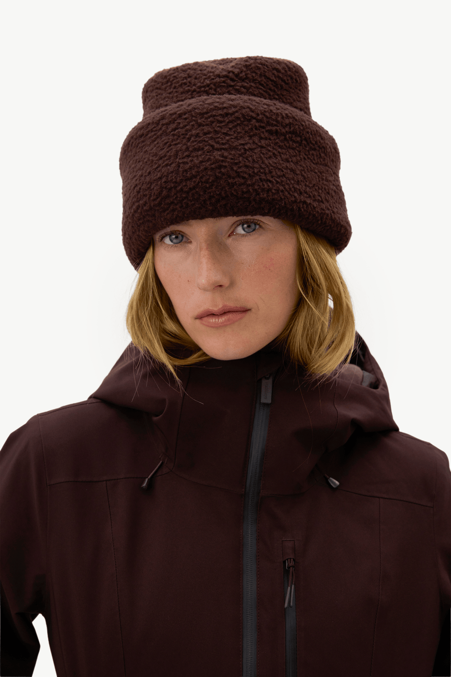 A woman wearing a dark brown fuzzy fleece beanie and a matching dark brown hooded jacket, photographed against a clean off-white studio background.