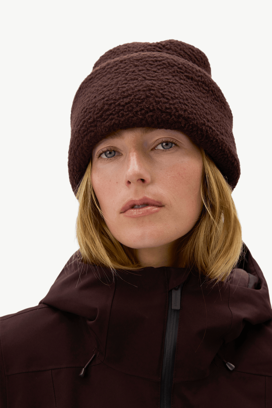 A close-up of a woman wearing a dark brown fuzzy fleece beanie and a matching dark brown hooded jacket against a plain light background.