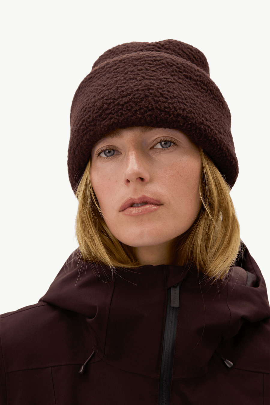 A close-up of a woman wearing a dark brown fuzzy fleece beanie and a matching dark brown hooded jacket against a plain light background.