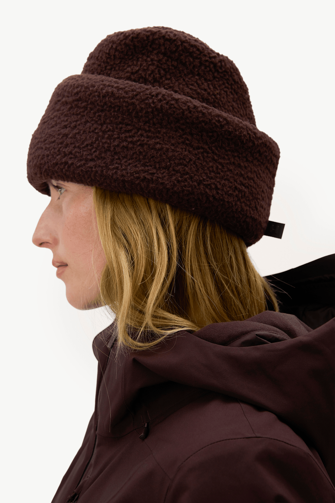 A woman in profile wearing a dark brown fuzzy fleece beanie and a matching dark brown hooded jacket against a plain white background.