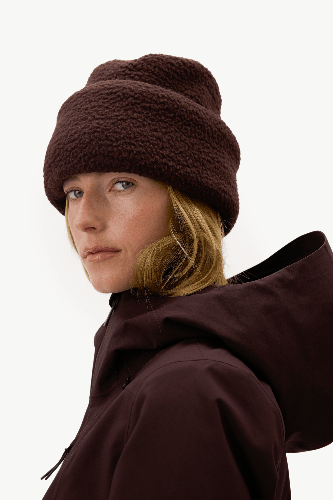 A woman wearing a dark brown textured fleece beanie and a matching dark brown hooded jacket, photographed against a clean off-white studio background.