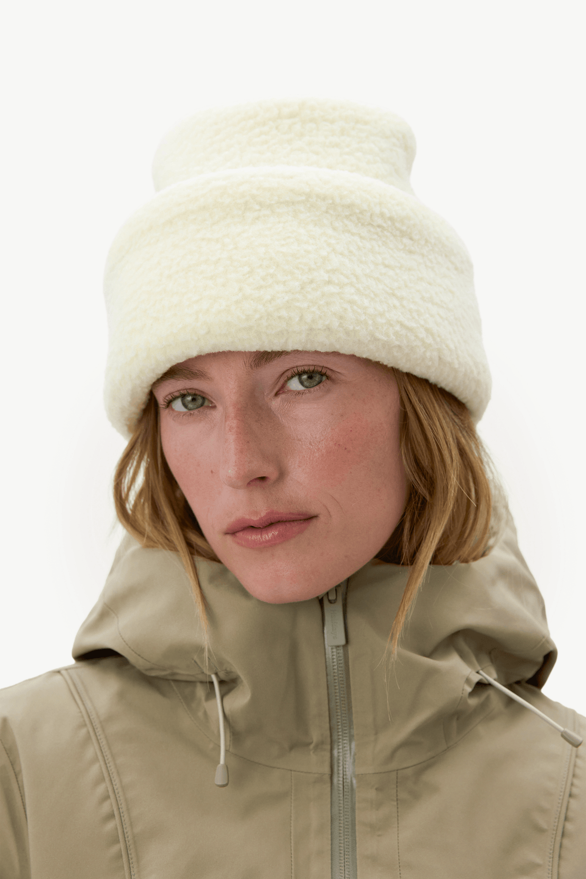 A close-up of a woman wearing a cream-colored, plush fleece beanie and a beige hooded jacket against a plain white background.