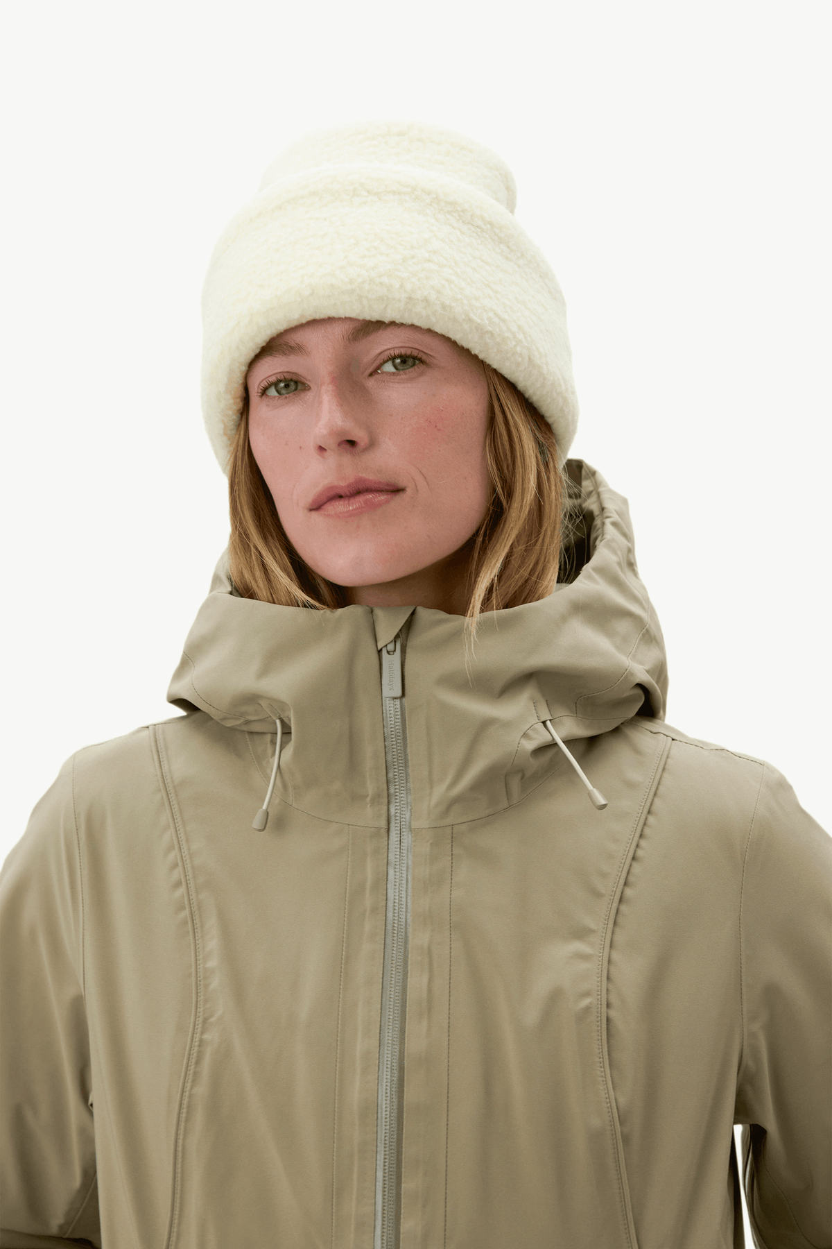 A model wearing a beige hooded ski jacket and a cream-colored fuzzy beanie hat, shown from the chest up against a plain white studio background.