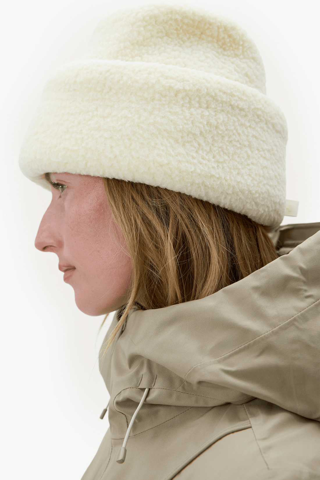 A close-up side profile of a woman wearing a thick, fluffy cream-colored beanie and a beige hooded jacket against a plain white background.