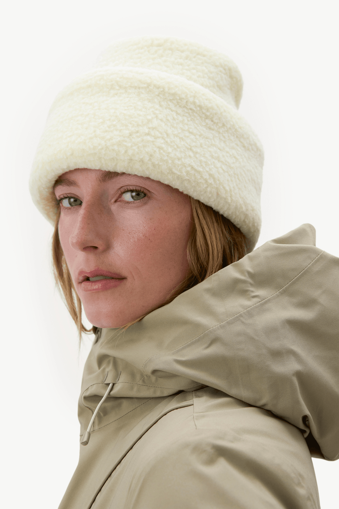 A close-up of a woman wearing a cream-colored, textured fleece beanie and a light beige hooded jacket against a plain white background.