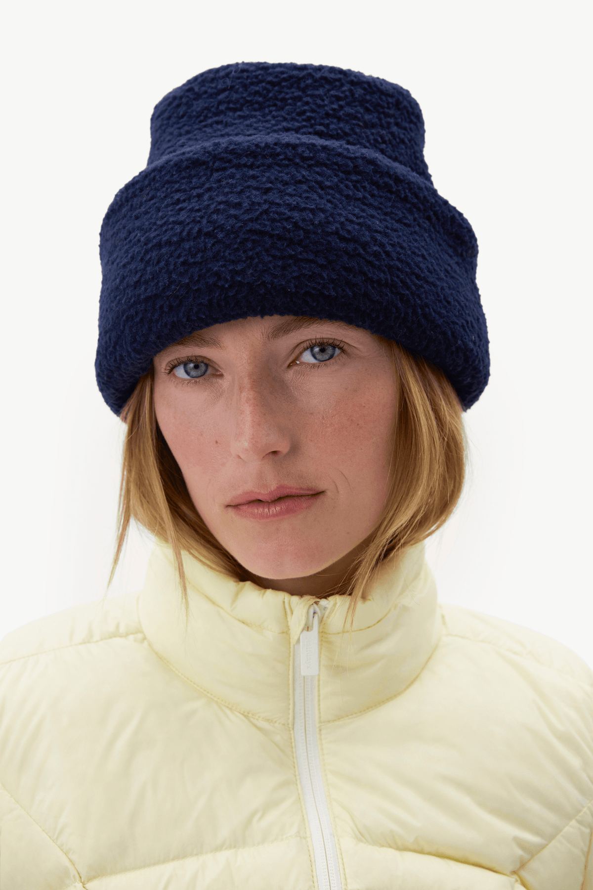 A woman wearing a dark navy blue fuzzy fleece beanie and a pale yellow puffer jacket, photographed against a clean white background.