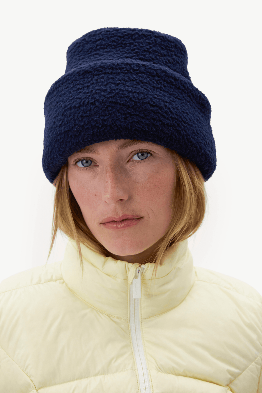 A woman wearing a dark navy blue fuzzy fleece beanie and a pale yellow puffer jacket, photographed against a clean white background.